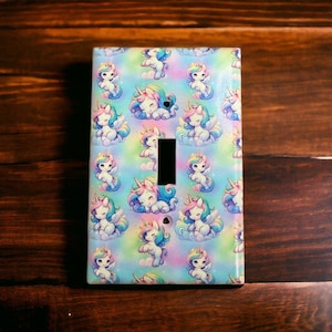 May include: A light switch cover with a colorful pattern of unicorns on a pastel blue background. The unicorns are white with pink and blue manes and tails.
