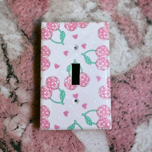 May include: A light switch cover with a pink and green design featuring cherries with a disco ball pattern. The background is a light pink with white glitter.