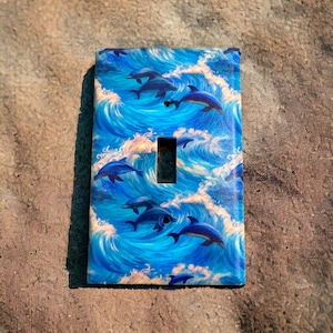 May include: A light switch cover with a blue and white design featuring dolphins swimming in waves.
