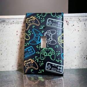 May include: A black light switch cover with a pattern of neon green, blue, and yellow video game controllers and buttons.