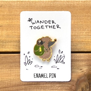 May include: An enamel pin featuring a cartoon hedgehog with a backpack, set against a white card with the words "Wander Together" and "Enamel Pin". The pin has gold trim and is on a wooden surface.