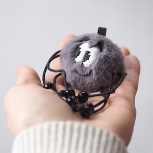 Puede incluir: Un personaje de pompón gris afelpado con ojos blancos bordados y una boca sonriente. Se adjuntan patas de cuerda negra y un lazo. Un artículo caprichoso hecho a mano.