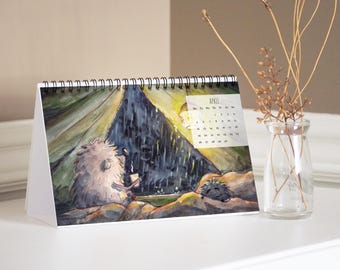 May include: A spiral-bound desk calendar featuring a watercolor illustration of two hedgehogs inside a tent during a rainstorm. The page displays the month of April with a grid of dates. The calendar is white with a black wire binding at the top.