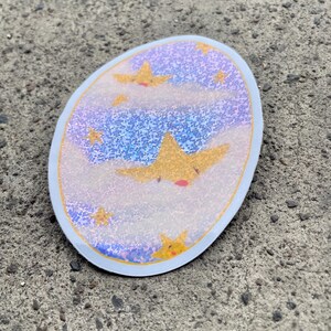 May include: A round sticker with a blue and purple glittery background with white clouds and yellow stars. The stars have a cartoon-like face with a red mouth.