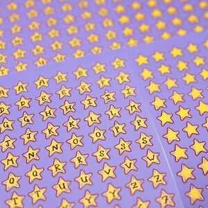 May include: Sheets of star-shaped stickers with letters and numbers on a purple background. The stickers have yellow stars with black outlines and black lettering. The letters A through Z and numbers 1 through 7 are visible.