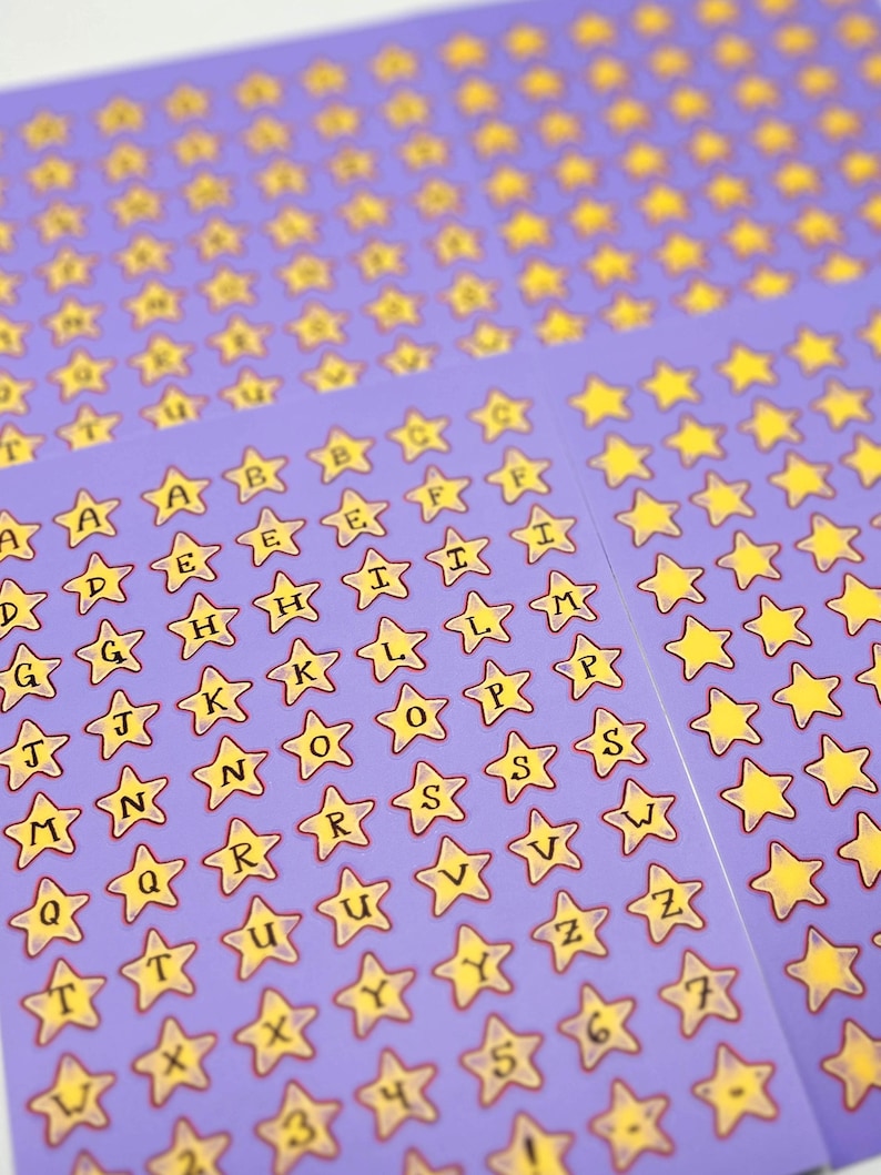 May include: Sheets of star-shaped stickers with letters and numbers on a purple background. The stickers have yellow stars with black outlines and black lettering. The letters A through Z and numbers 1 through 7 are visible.