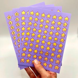May include: Sheets of purple stickers featuring yellow star designs with various facial expressions. Some sheets have the letters A, D, G, J, and M. The stickers are held in a hand against a white background.