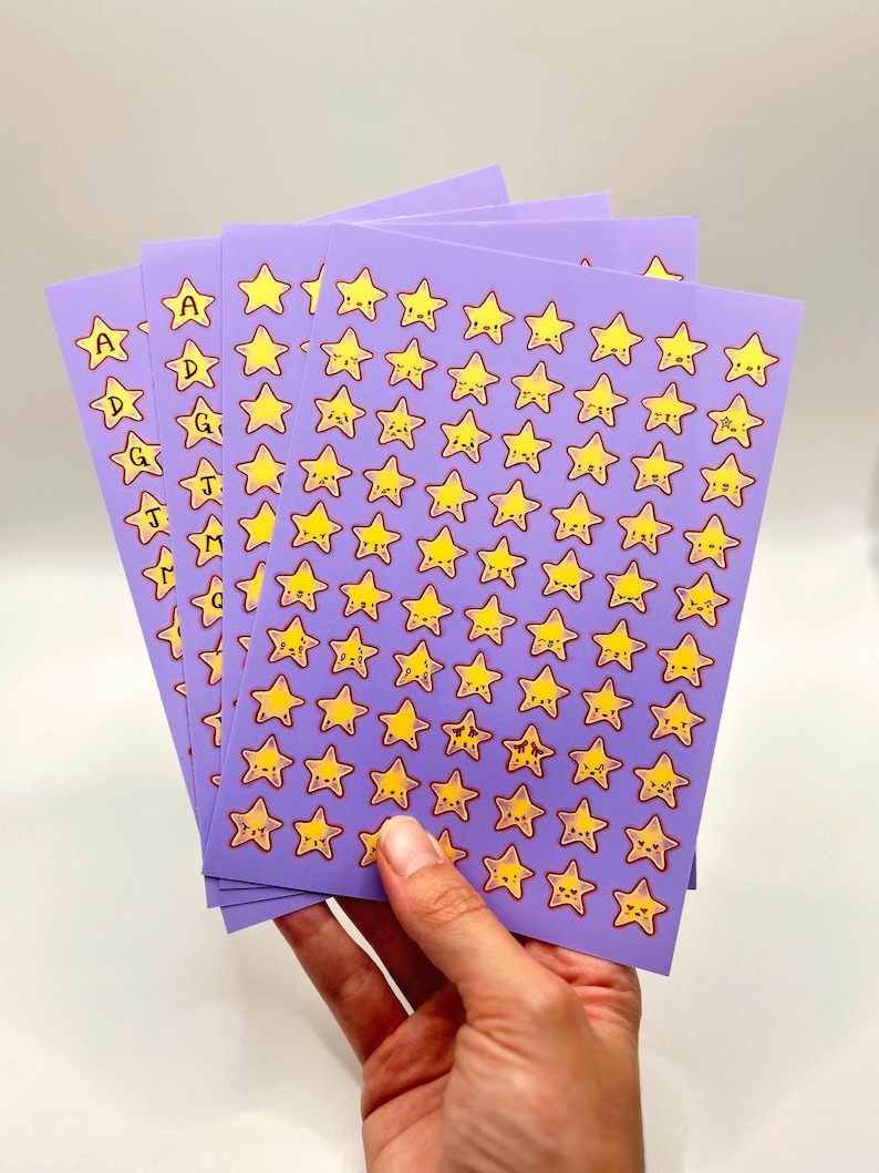 May include: Sheets of purple stickers featuring yellow star designs with various facial expressions. Some sheets have the letters A, D, G, J, and M. The stickers are held in a hand against a white background.