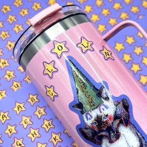 May include: A pink insulated tumbler with a handle and a lid. The tumbler is decorated with star stickers and a sticker of a cat wearing a dunce cap. The word "DUNCE" is visible on the cap.
