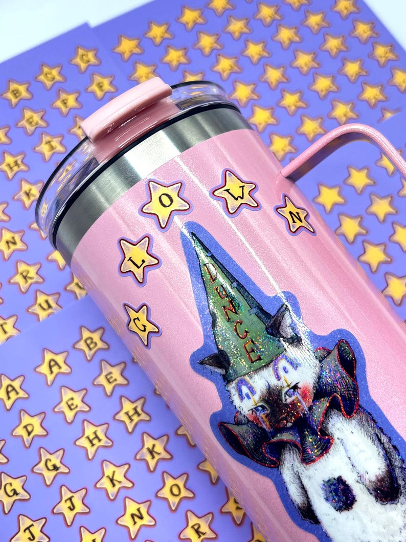 May include: A pink insulated tumbler with a handle and a lid. The tumbler is decorated with star stickers and a sticker of a cat wearing a dunce cap. The word "DUNCE" is visible on the cap.
