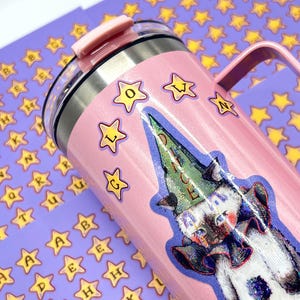 May include: A pink insulated tumbler with a handle and a clear lid. The tumbler is decorated with star stickers and a cartoon cat wearing a wizard hat. The word "LOVE" is spelled out with star stickers. The background features star and letter stickers.