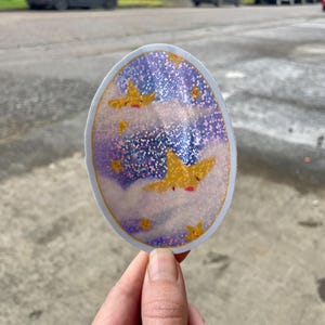 May include: A round sticker with a glittery purple and blue background. The sticker features a white cloud with a yellow star with a face on it. There are other yellow stars scattered throughout the sticker.
