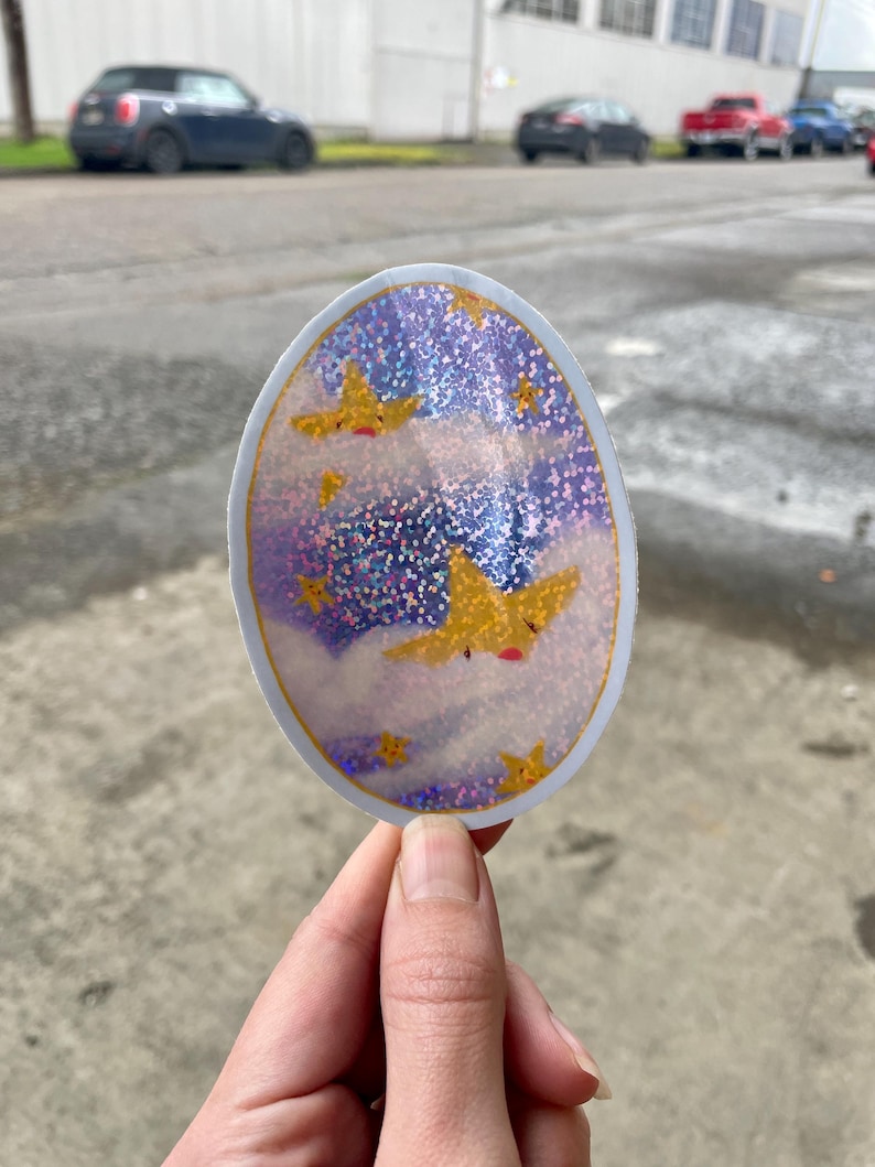 May include: A round sticker with a glittery purple and blue background. The sticker features a white cloud with a yellow star with a face on it. There are other yellow stars scattered throughout the sticker.