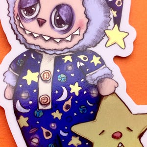 May include: A blue and purple fuzzy monster wearing a nightcap and pajamas with a space theme print. The monster is holding a yellow star with a sleepy face.