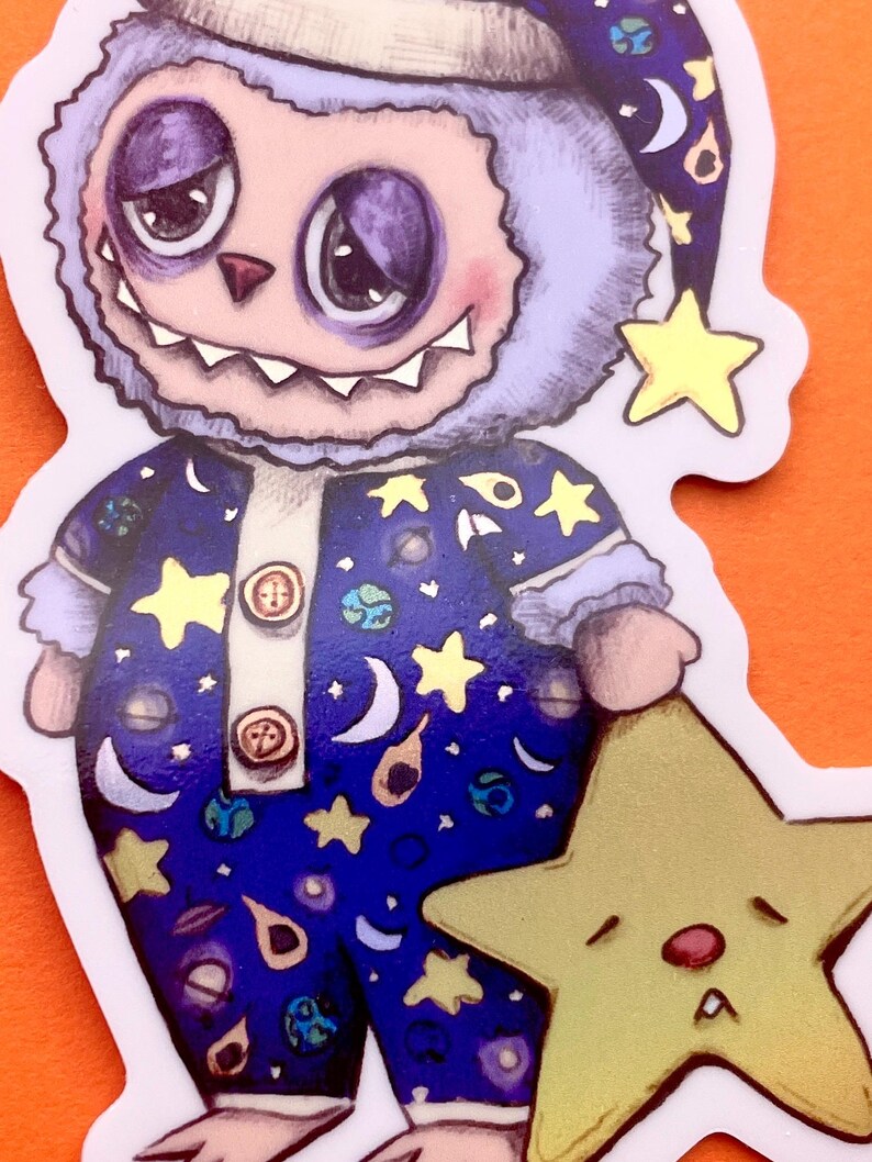May include: A blue and purple fuzzy monster wearing a nightcap and pajamas with a space theme print. The monster is holding a yellow star with a sleepy face.