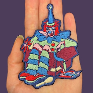 May include: An embroidered iron-on patch featuring a sad clown sitting in a chair. The clown is wearing a blue and green costume with red pom-poms and a blue hat with a star. The clown has red hair and a blue face with a tear.