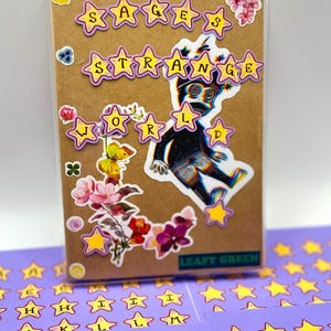 May include: A brown notebook with the words "SAGES STRANGE WORLD" in star-shaped stickers. The notebook is decorated with floral stickers and a sticker of a figure. Additional star-shaped letter stickers are visible.