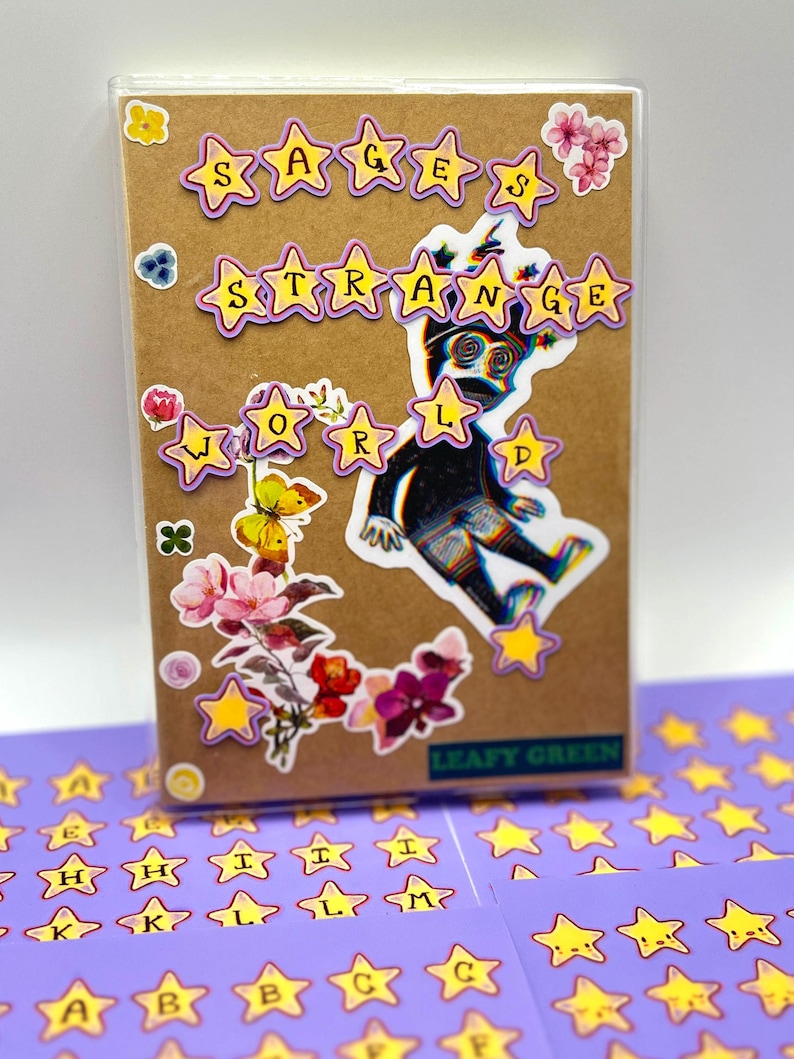 May include: A brown notebook with the words "SAGES STRANGE WORLD" in star-shaped stickers. The notebook is decorated with floral stickers and a sticker of a figure. Additional star-shaped letter stickers are visible.