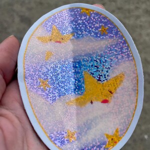May include: A round sticker with a blue background and a holographic finish. The sticker features a design of yellow stars with faces on a white cloud background.
