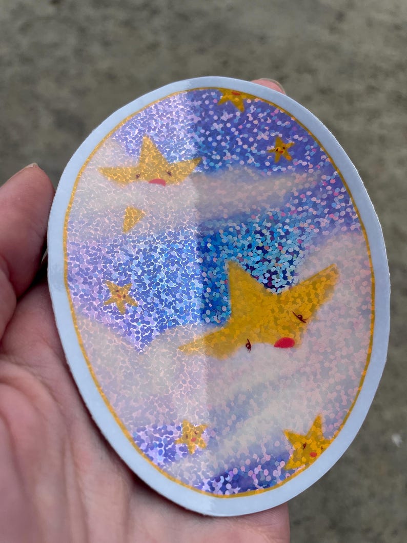 May include: A round sticker with a blue background and a holographic finish. The sticker features a design of yellow stars with faces on a white cloud background.