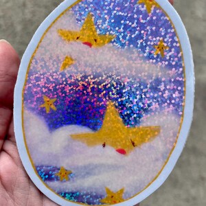 May include: A round sticker with a holographic background of blue, purple, and pink glitter. The sticker features a white cloud with a yellow star with a red mouth and closed eyes. There are other yellow stars with red mouths and closed eyes on the sticker.
