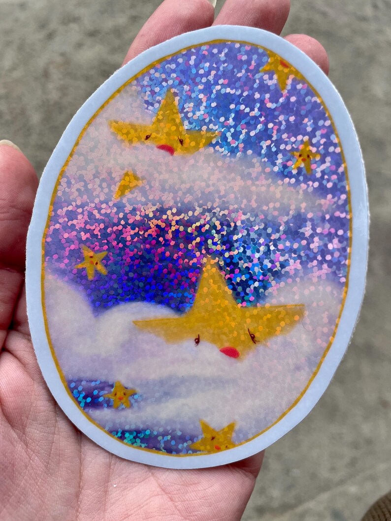 May include: A round sticker with a holographic background of blue, purple, and pink glitter. The sticker features a white cloud with a yellow star with a red mouth and closed eyes. There are other yellow stars with red mouths and closed eyes on the sticker.