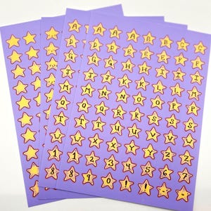 May include: Sheets of purple star-shaped stickers with yellow stars and black lettering. Each sticker features a letter of the alphabet or a number. The stickers are arranged in rows and columns, with a variety of letters and numbers.