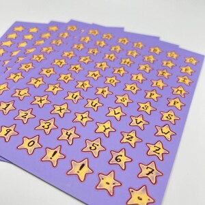 May include: Sheets of star-shaped stickers with letters, numbers, and symbols. The stickers have a yellow star design with a red outline, set against a light purple background. The letters and numbers are in black.
