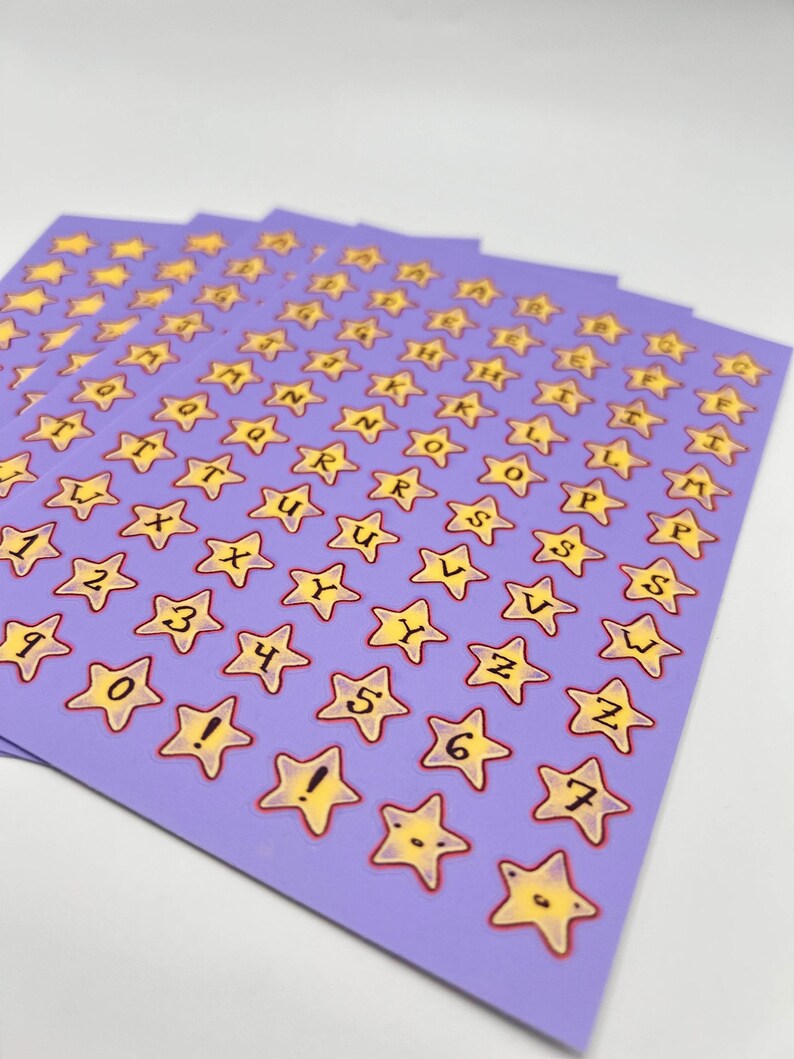 May include: Sheets of star-shaped stickers with letters, numbers, and symbols. The stickers have a yellow star design with a red outline, set against a light purple background. The letters and numbers are in black.