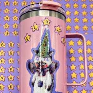 May include: A pink insulated tumbler with a handle, featuring a cartoon cat wearing a dunce cap. The tumbler has a silver rim and a pink lid. The background is covered in star stickers and alphabet stickers.