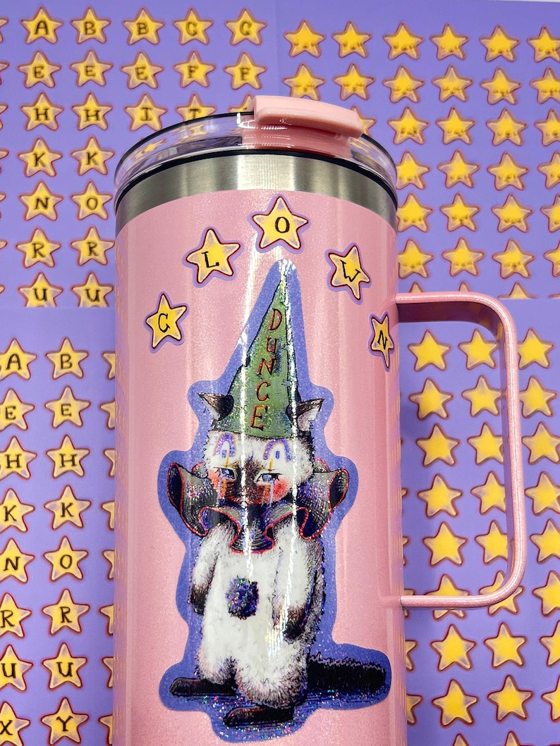 May include: A pink insulated tumbler with a handle, featuring a cartoon cat wearing a dunce cap. The tumbler has a silver rim and a pink lid. The background is covered in star stickers and alphabet stickers.