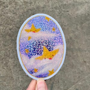May include: A round sticker with a blue and purple glitter background. The sticker features three yellow stars with red faces, flying through white clouds.