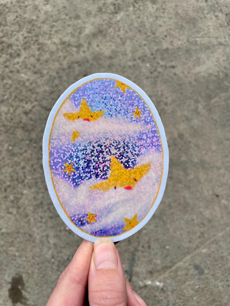 May include: A round sticker with a blue and purple glitter background. The sticker features three yellow stars with red faces, flying through white clouds.
