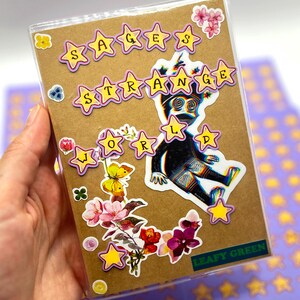 May include: A brown notebook with the words "Sages Strange World" in yellow star-shaped stickers. The notebook is decorated with a black and white illustration of a creature, along with floral and butterfly stickers. The words "Leafy Green" are also visible.