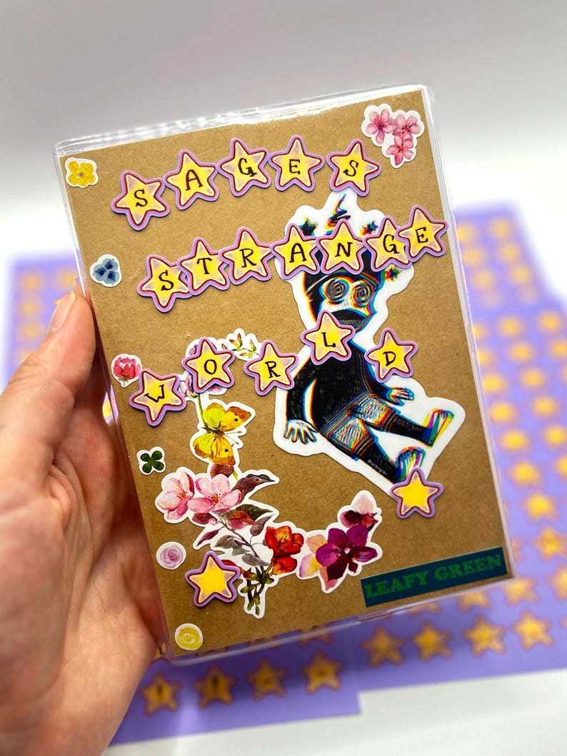 May include: A brown notebook with the words "Sages Strange World" in yellow star-shaped stickers. The notebook is decorated with a black and white illustration of a creature, along with floral and butterfly stickers. The words "Leafy Green" are also visible.
