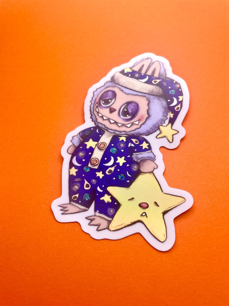 May include: A purple and blue fuzzy monster wearing a blue and white nightcap with stars and planets printed on it. The monster is holding a yellow star with a sleepy face.
