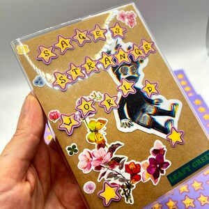 May include: A clear plastic case with a brown card featuring stickers. The stickers include yellow stars, a black figure, and floral designs. Text on the card reads "SAGES STRANGE WORLD" and "LEAFY GREEN".