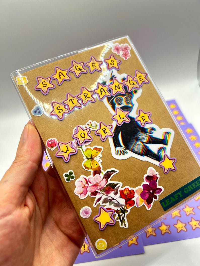 May include: A clear plastic case with a brown card featuring stickers. The stickers include yellow stars, a black figure, and floral designs. Text on the card reads "SAGES STRANGE WORLD" and "LEAFY GREEN".