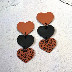 May include: A pair of dangle earrings with three heart-shaped charms. The top charm is solid orange, the middle charm is black, and the bottom charm is orange with a black leopard print.