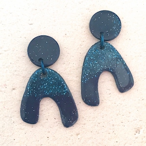 Op de afbeelding: Een paar donkerblauwe en turquoise glitter statement oorbellen met een unieke boogvorm.