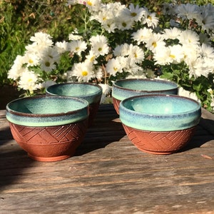 May include: Four terracotta bowls with a teal glaze and a carved pattern around the rim. The bowls are sitting on a wooden table.