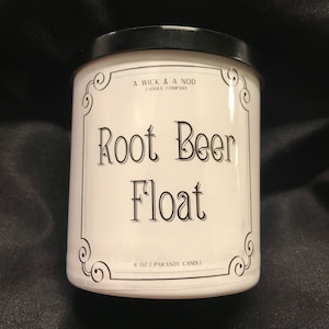 Puede incluir: Un tarro de vela blanco con tapa negra, con el texto "Root Beer Float" en escritura elegante. La etiqueta incluye el texto "A Wick & A Nod Candle Company" y "8 oz | Parasoy Candle". El tarro tiene adornos decorativos en las esquinas.