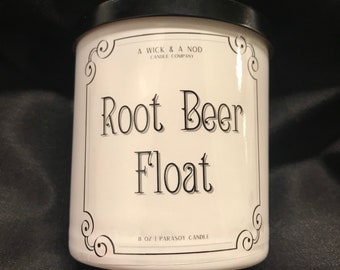 Peut inclure: Un pot de bougie blanc avec un couvercle noir, avec le texte "Root Beer Float" en écriture élégante. L'étiquette comprend le texte "A Wick & A Nod Candle Company" et "8 oz | Parasoy Candle". Le pot a des ornements décoratifs aux coins.