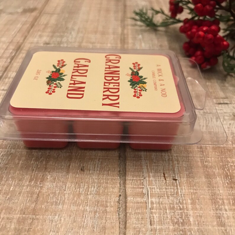 May include: A clear plastic clamshell package containing six red wax melts. The label reads "Cranberry Garland" with floral accents. The package is from "A Wick & A Nod Candle Company". The net weight is 2.65 oz.