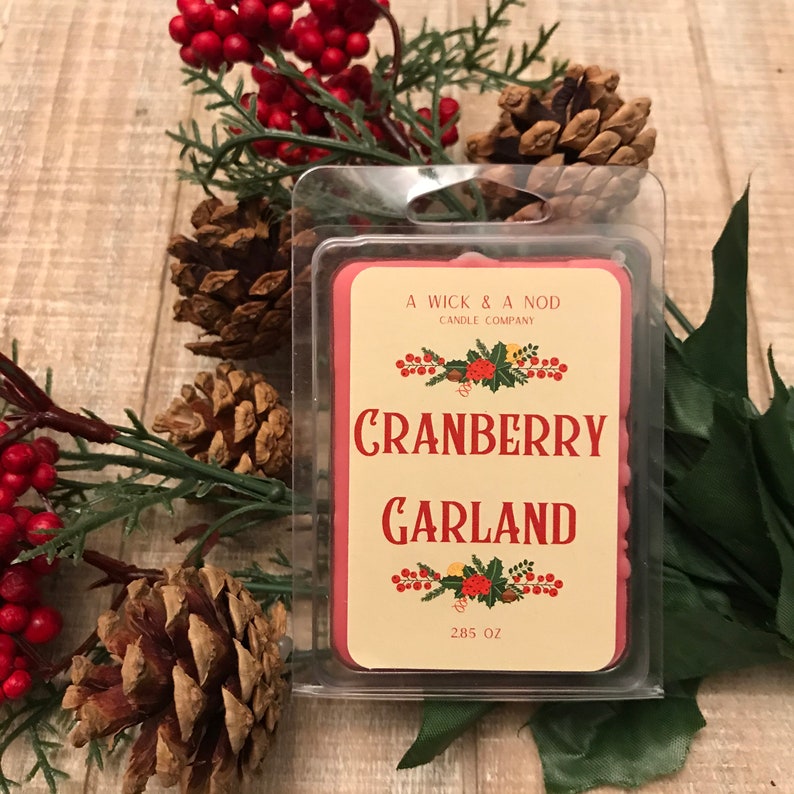 May include: A Wick & Nod Candle Company cranberry garland wax melt in a clear plastic container. The label is cream-colored with red text and a cranberry and holly design. The wax melt is surrounded by pine cones, red berries, and greenery.