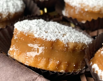 Op de afbeelding: Close-up van een doos met kleine, ronde, met karamel gevulde gebakjes die bestrooid zijn met poedersuiker. De gebakjes zijn gerangschikt in een doos die is bekleed met bruine papieren strooisel. De tekst "MANJAR BY SANDRA" is zichtbaar in de rechterbenedenhoek van de afbeelding.
