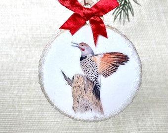 Könnte beinhalten: Runder Weihnachtsbaumschmuck aus Holz mit einer detaillierten Illustration eines Vogels, der auf einem Baumstumpf sitzt. Der Schmuck hat einen glitzernden Rand, eine rote Satinschleife und eine silberne Aufhängeschlaufe. Eine festliche Weihnachtsdekoration.