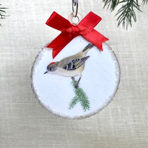 May include: Round Christmas ornament featuring a detailed illustration of a small bird perched on a pine branch. The ornament has a white background with a silver glitter border and a red satin bow. A metal clip is attached for hanging.