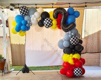 May include: A balloon arch made of black, white, red, yellow, blue, and gray balloons. The arch is decorated with checkered balloons and a large black number two balloon. The arch is set up in front of a white backdrop.