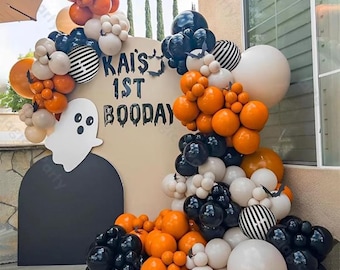 May include: A Halloween-themed balloon arch with orange, black, white, and striped balloons. The arch surrounds a sign that reads "Kai's 1st Booday" with a ghost cutout. The balloons are arranged in clusters and arches, creating a festive party decoration.